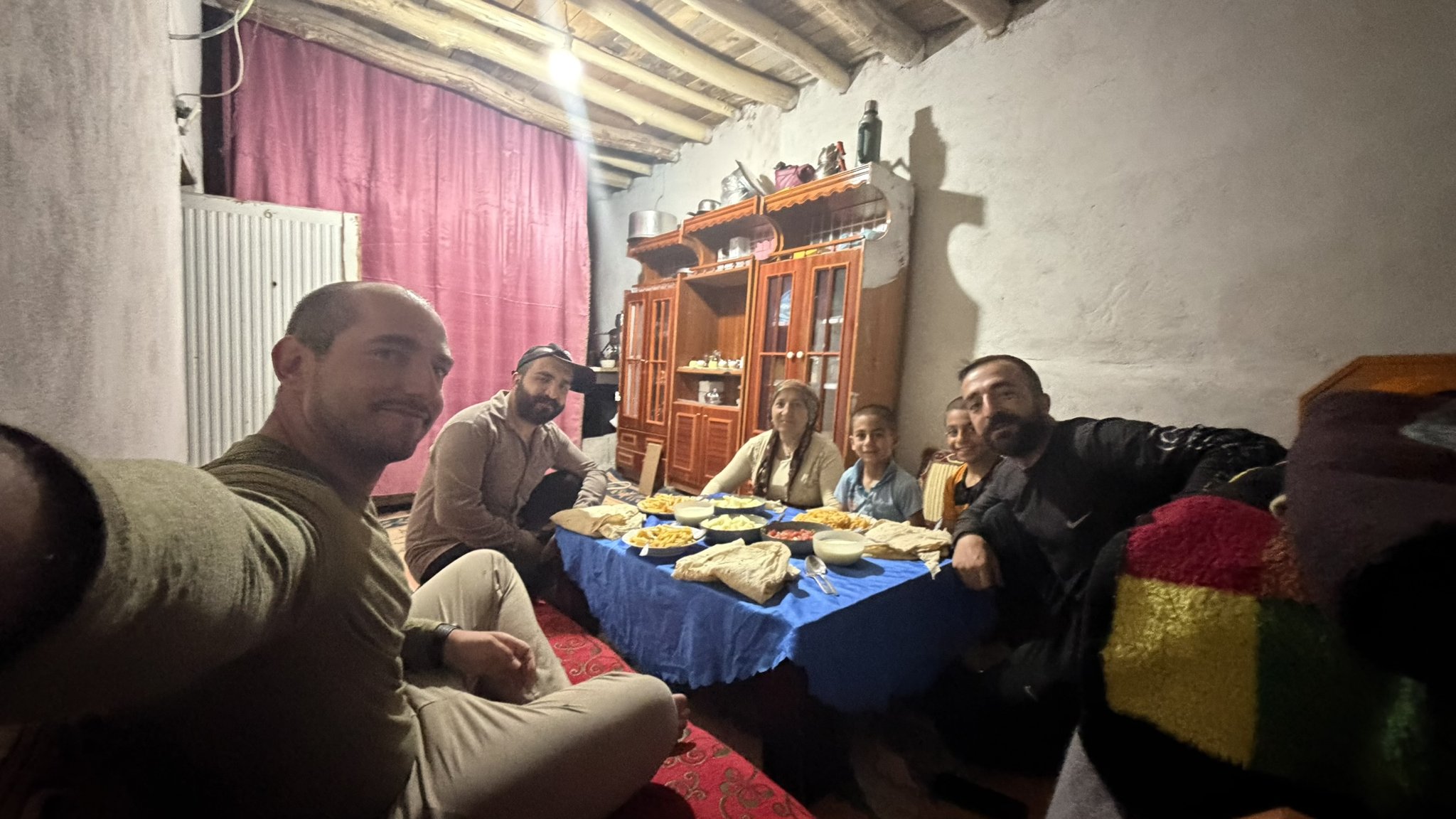 Gruppe von Menschen sitzt am Boden um einen gedeckten Tisch mit Fladenbrot und Beilagen in einem einfachen Wohnraum.