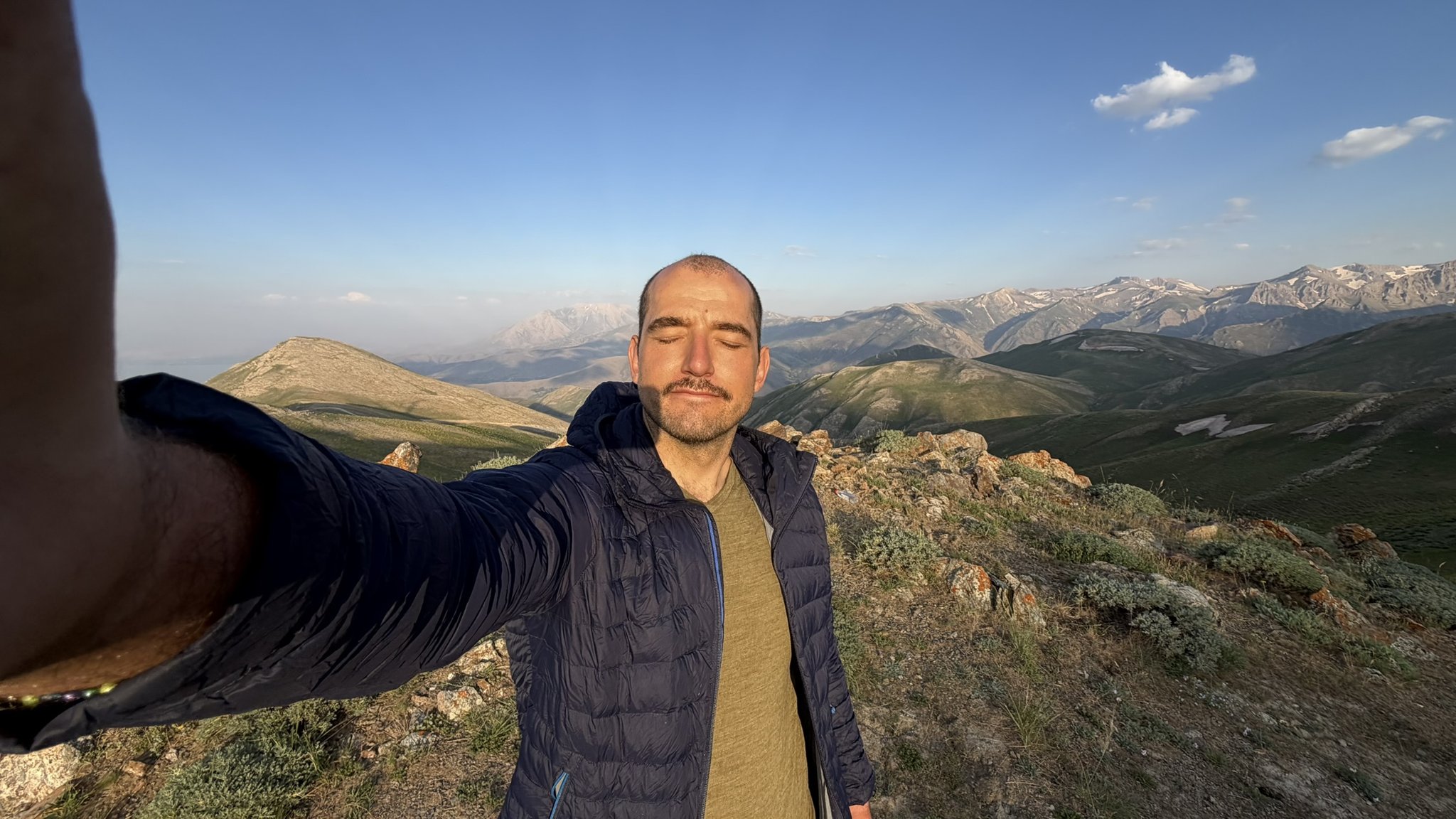 Selfie in Daunenjacke auf einem Bergrücken, im Hintergrund schneebedeckte Gipfel im Abendlicht.