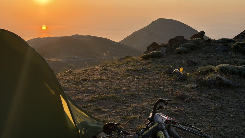Zelt und bepacktes Bikepacking-Rad im Morgenlicht auf einem Bergrücken in Ostanatolien, dahinter Sonnenaufgang über dem Dunst des Vansees.