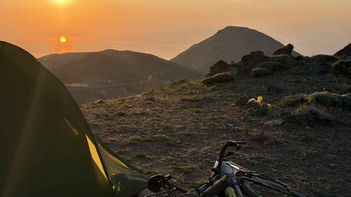Zelt und bepacktes Bikepacking-Rad im Morgenlicht auf einem Bergrücken in Ostanatolien, dahinter Sonnenaufgang über dem Dunst des Vansees.