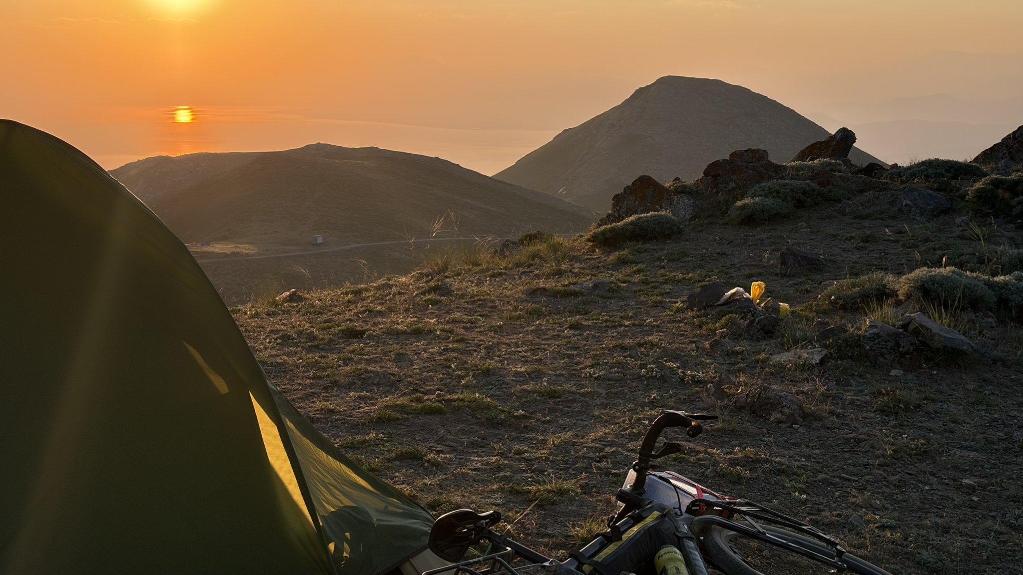 Zelt und bepacktes Bikepacking-Rad im Morgenlicht auf einem Bergrücken in Ostanatolien, dahinter Sonnenaufgang über dem Dunst des Vansees.