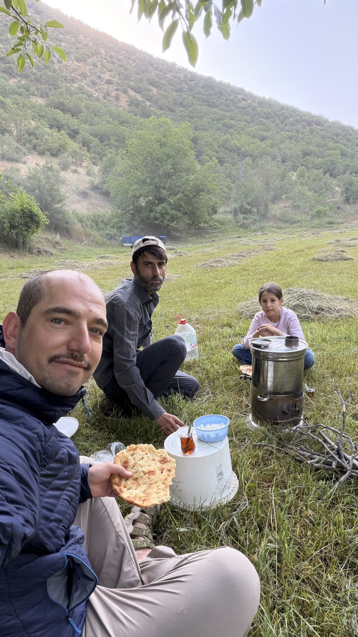 Moritz sitzt mit einem Mann und einem Mädchen auf einer Wiese, Fladenbrot in der Hand, Teekessel auf dem Boden.