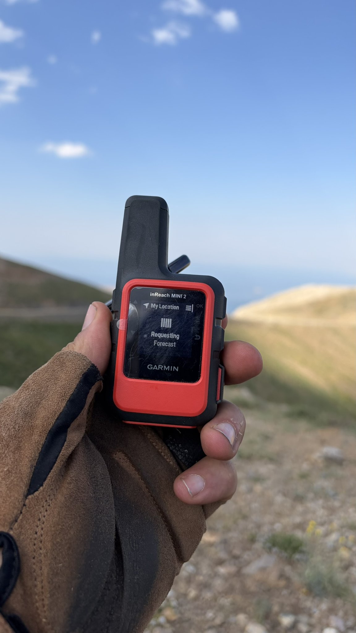 Hand mit Garmin inReach Mini 2 auf einem Bergpass, Display zeigt Requesting Forecast.